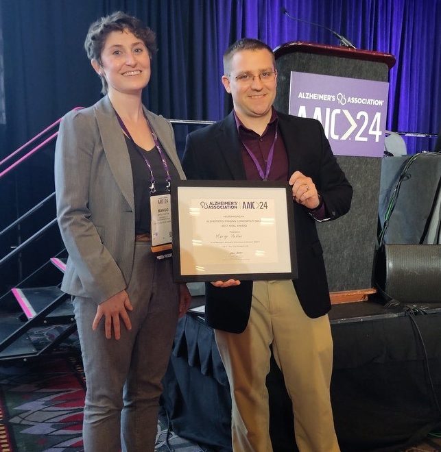 Margo Heston and Tobey Betthauser holding AIC Best Oral Presentation Award in front of AAIC Stage Podium.