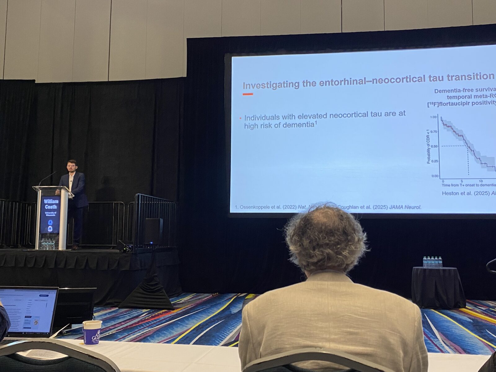 Postdoc Will Coath at the podium presenting at HAI 2026; research slide of talk shows in background.